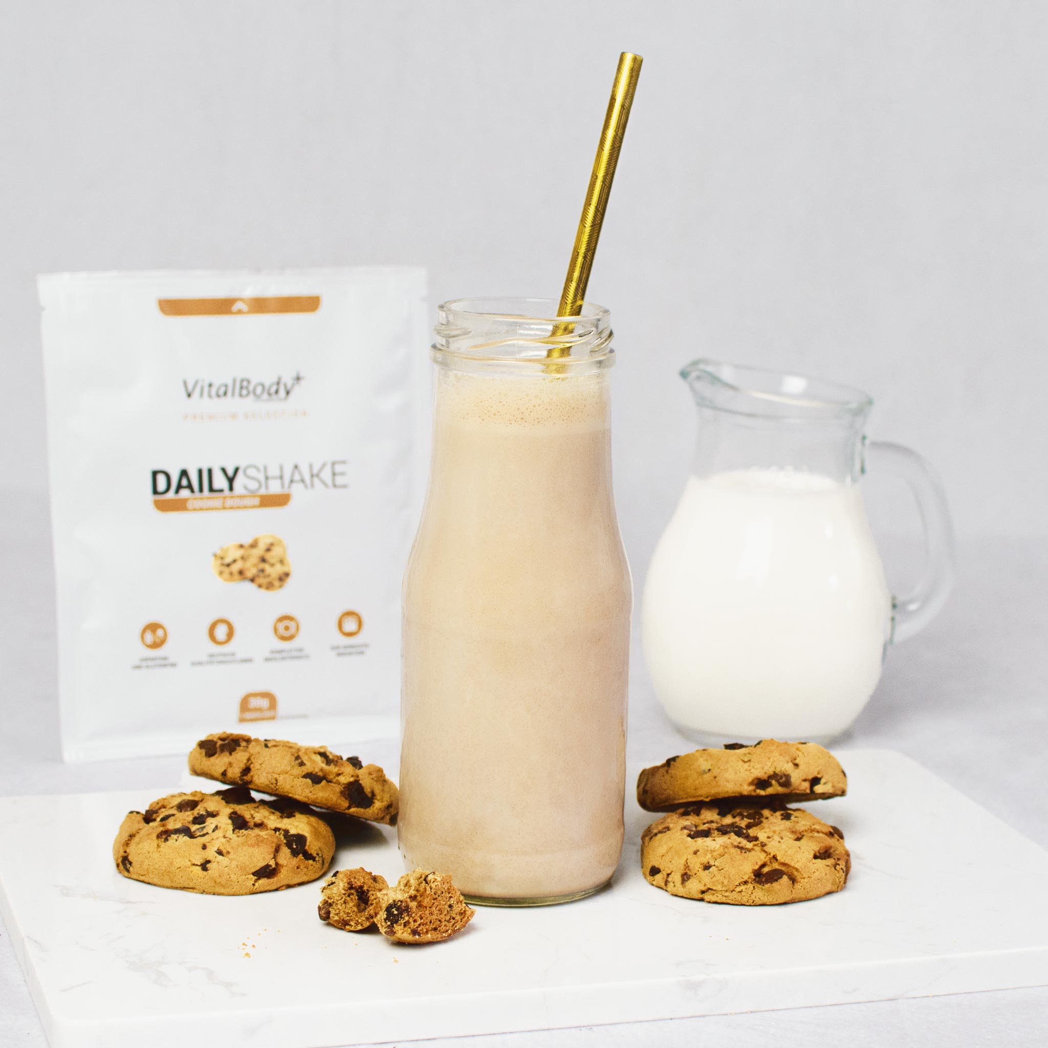 Glasflasche mit Strohalm und Shakeinhalt Cookie Dough mit Cookies auf einer Marmorplatte dahinter ein DailyShake To Go Pack Cookie Dough und eine Karaffe mit Hafermilch