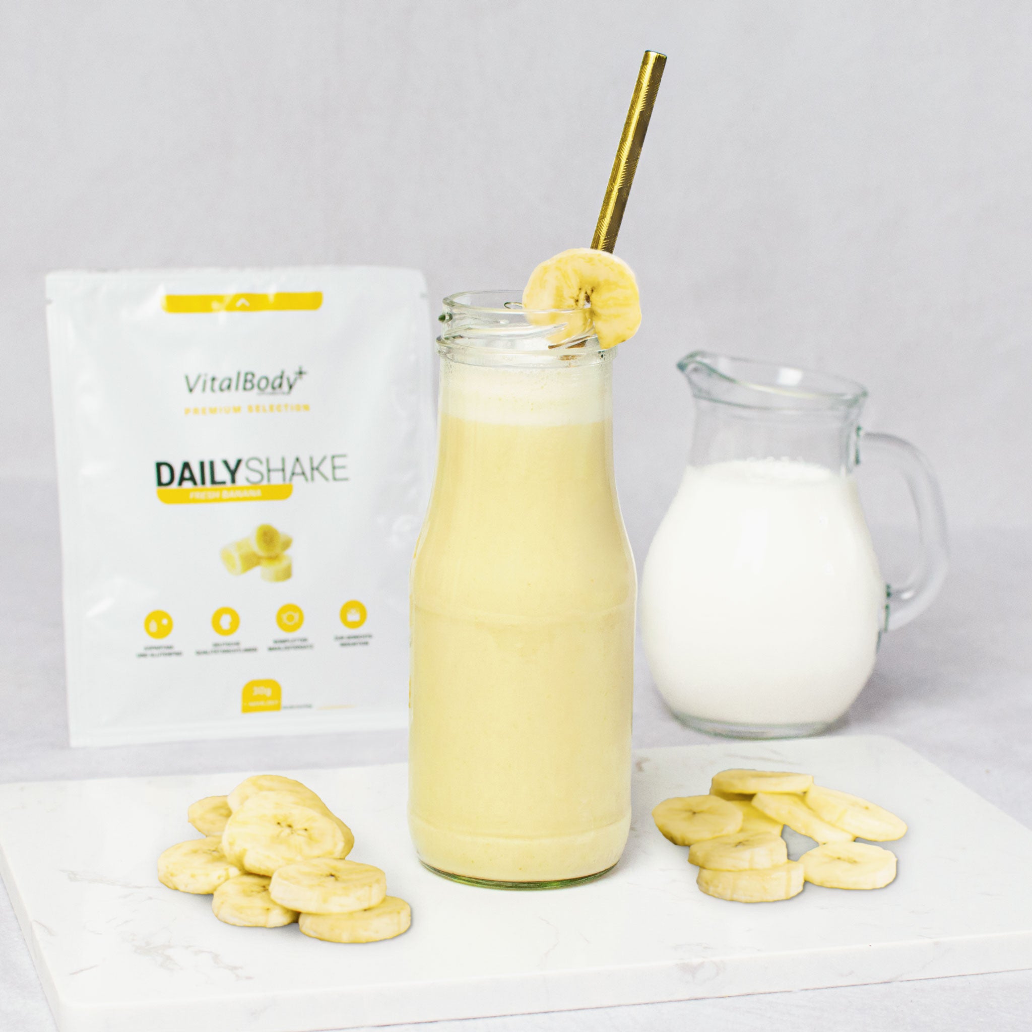 Glasflasche mit Shakeinhalt Fresh Banana mit Bananenscheiben auf einer Marmorplatte im Hintergrund ein DailyShake To Go Pack Fresh Banana und einer Karaffe mit Hafermilch