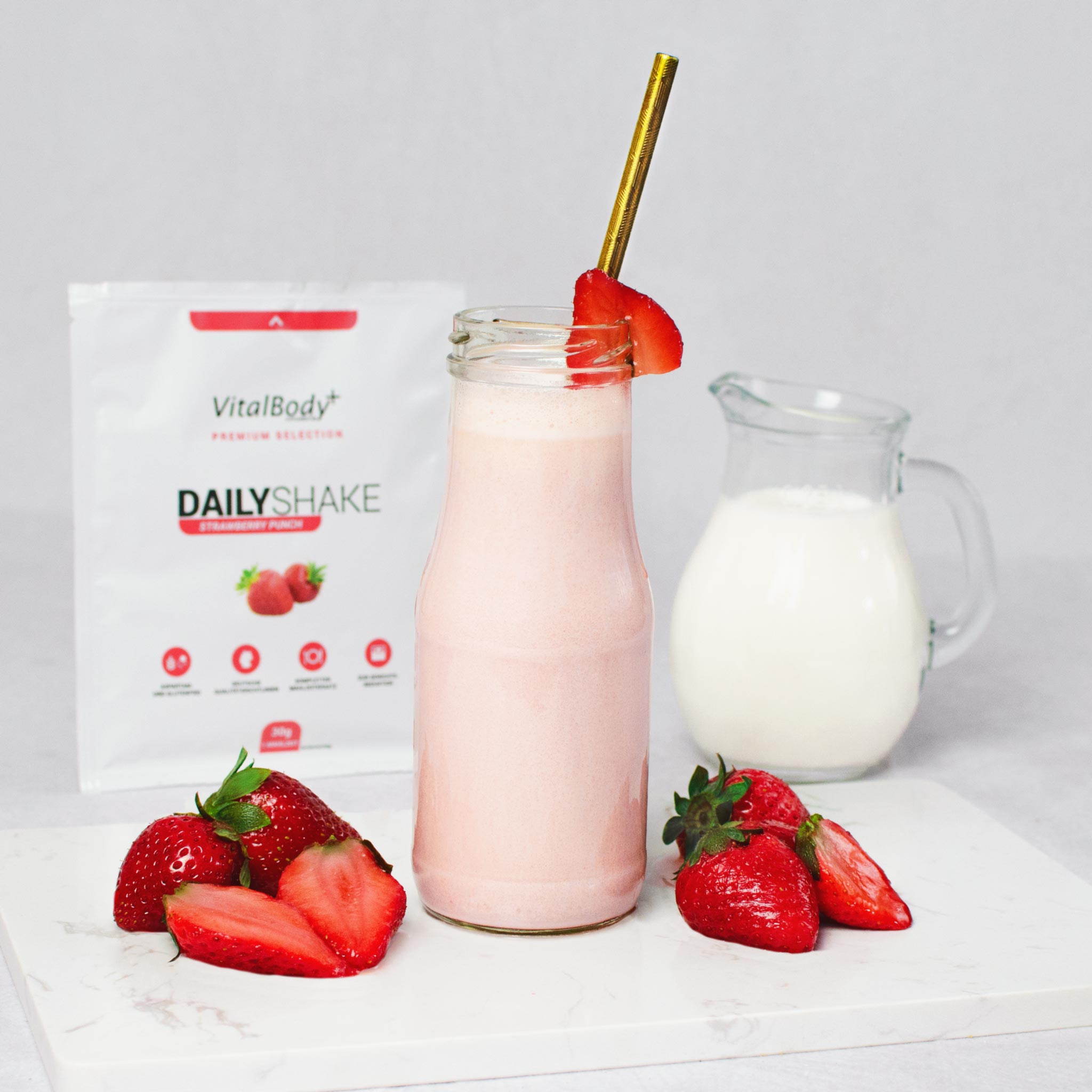 Glasflasche mit Shakeinhalt Strawberry Punch mit Erdbeeren auf einer Marmorplatte im Hintergrund ein DailyShake To Go Pack Strawberry Punch und einer Karaffe mit Hafermilch