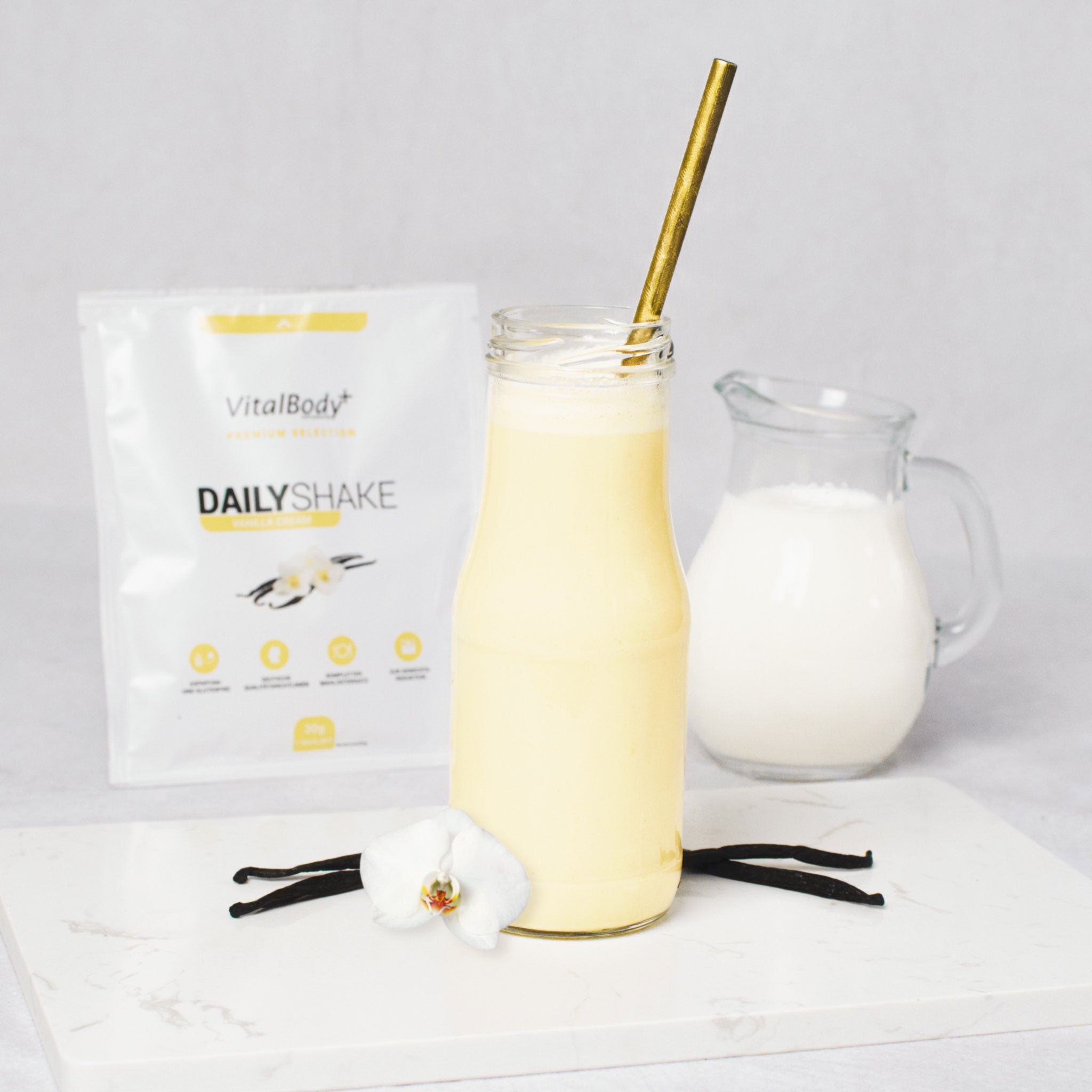 Glasflasche mit Shakeinhalt Vanilla Cream mit Vanilleschote und Blüte auf einer Marmorplatte im Hintergrund ein DailyShake To Go Pack Vanilla Cream und einer Karaffe mit Hafermilch