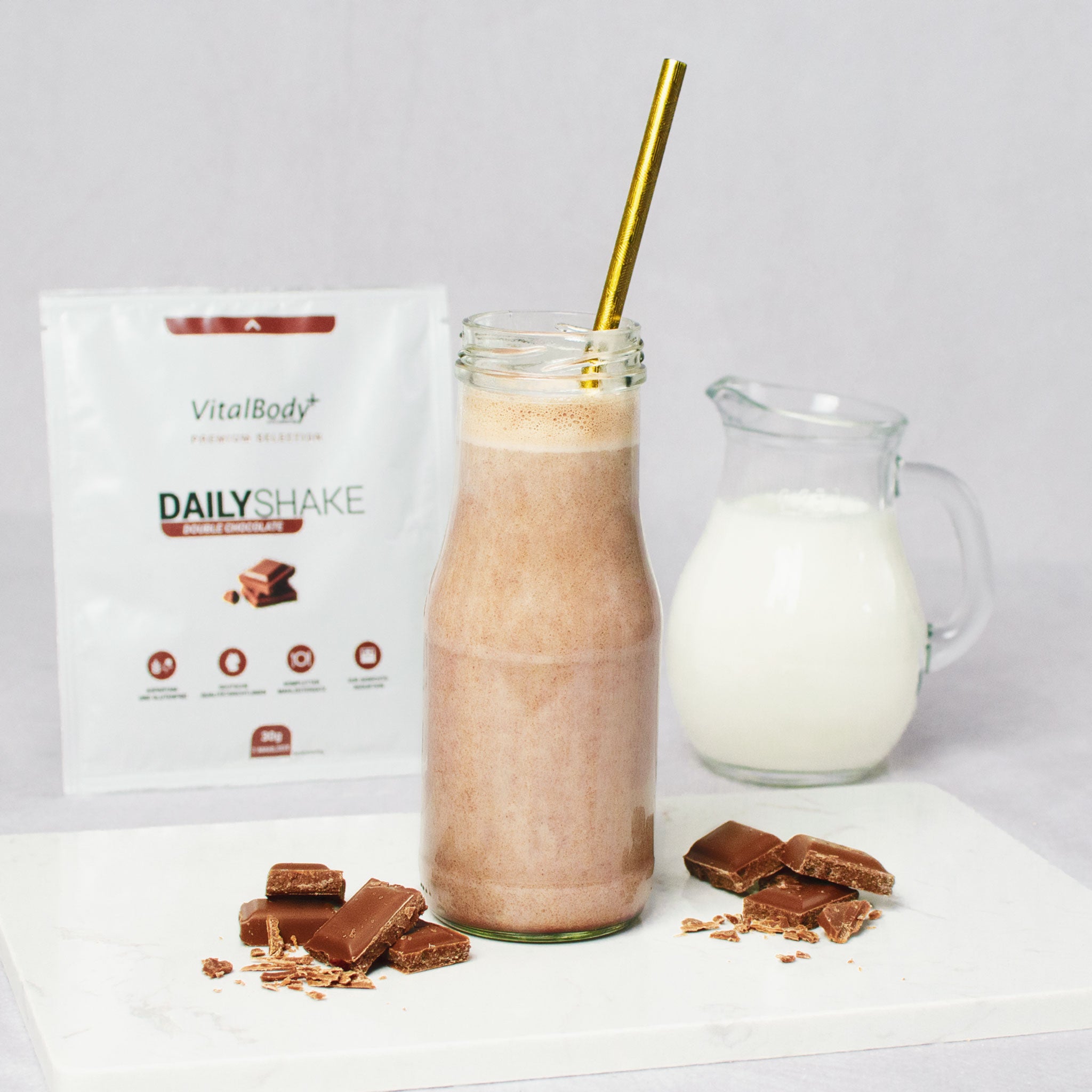 Glasflasche mit Shakeinhalt Double Chocolate mit Schokoladenstückchen auf einer Marmorplatte im Hintergrund ein DailyShake To Go Pack Double Chocolate und einer Karaffe mit Hafermilch