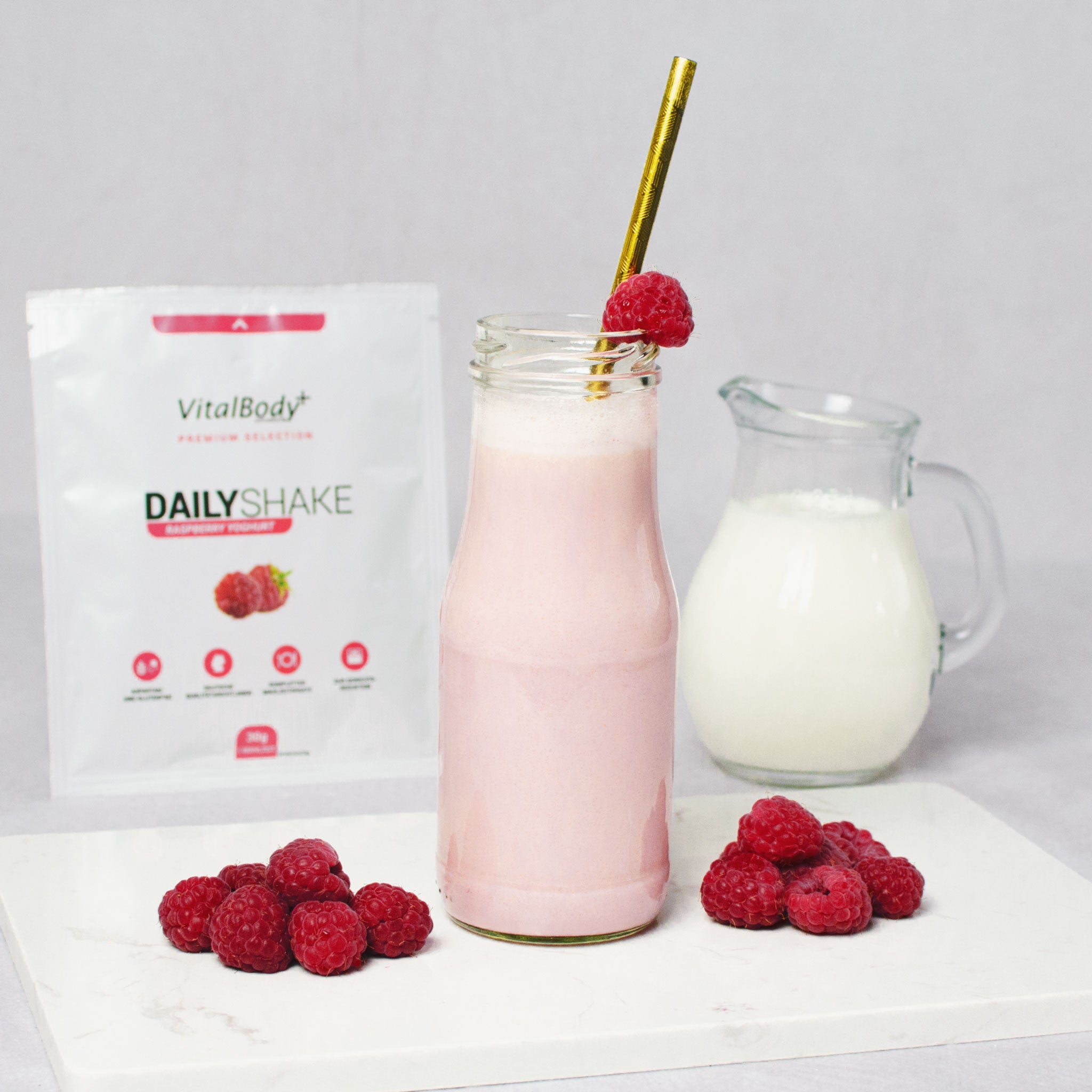 Glasflasche mit Shakeinhalt Raspberry Yoghurt mit Himbeeren auf einer Marmorplatte im Hintergrund ein DailyShake To Go Pack Raspberry Yoghurt und einer Karaffe mit Hafermilch